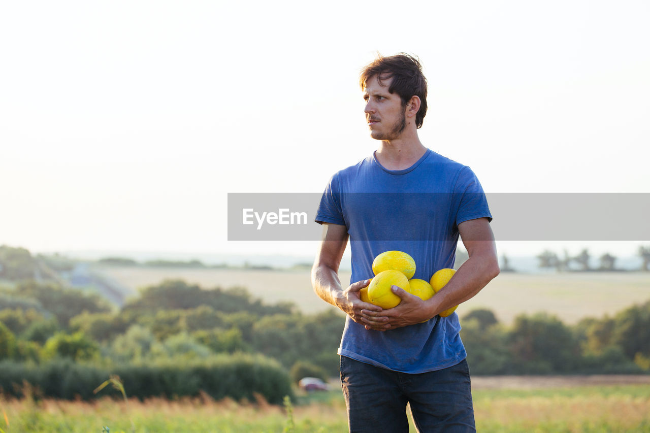 Farmer holding fruits while standing on land against clear sky