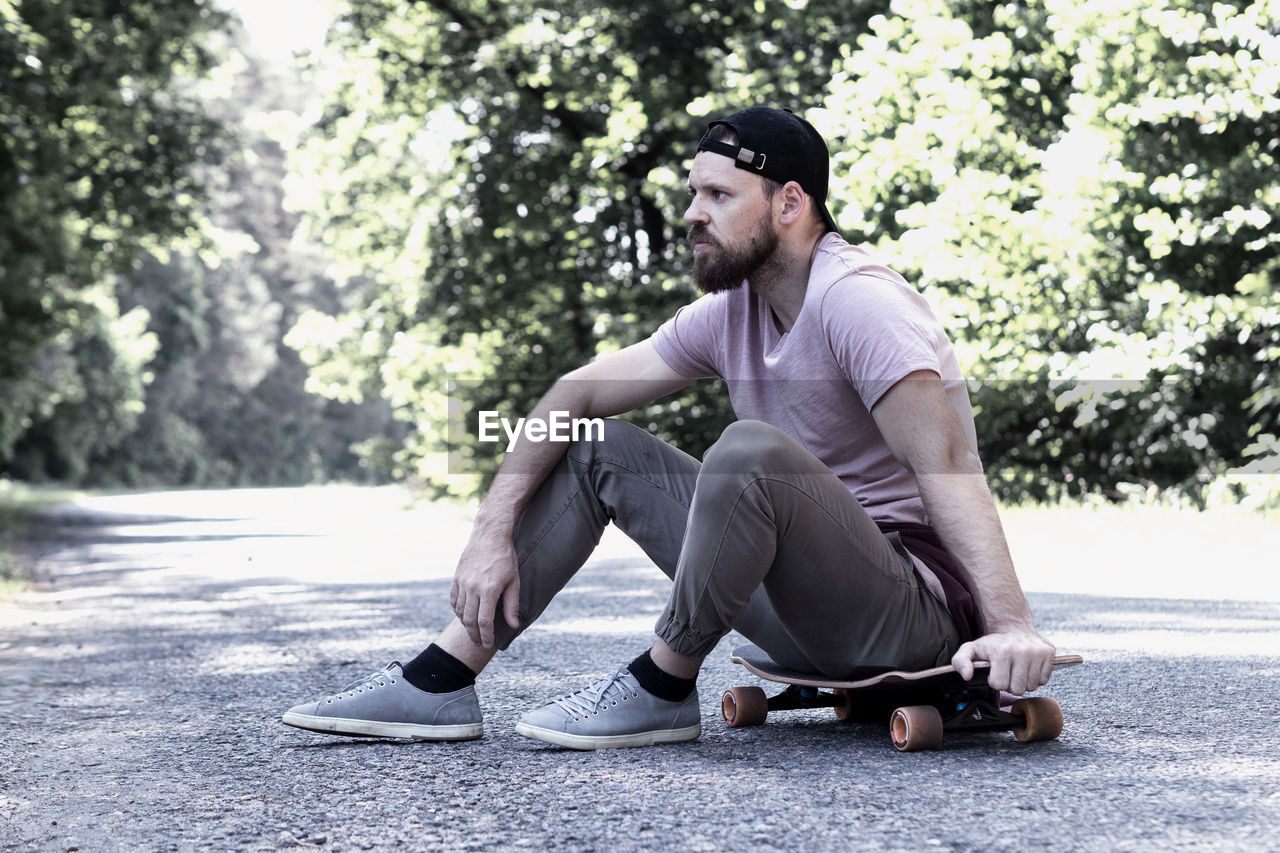 Thoughtful full length of man sitting on skateboard at road