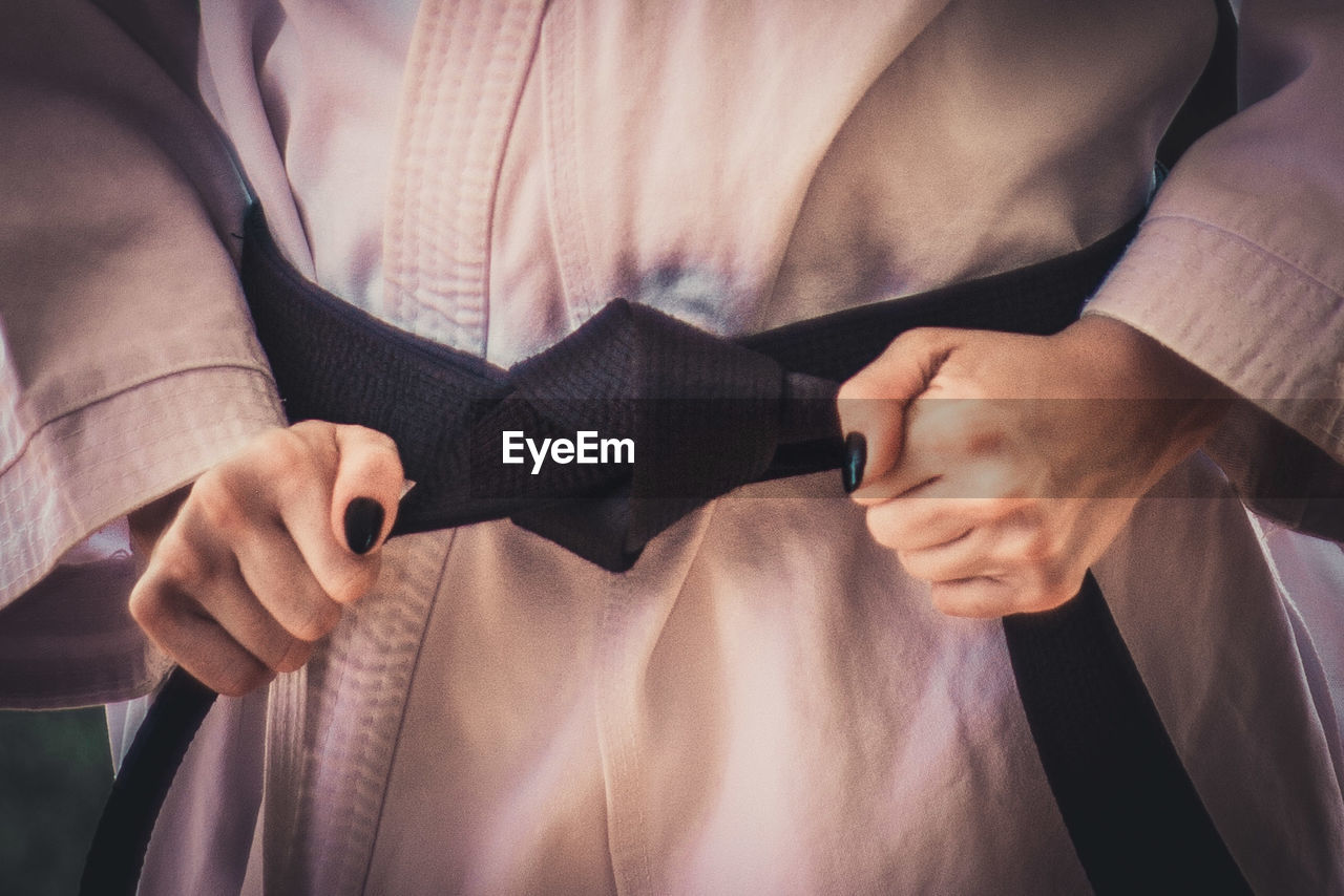 Midsection of woman tying black belt while standing in karate uniform