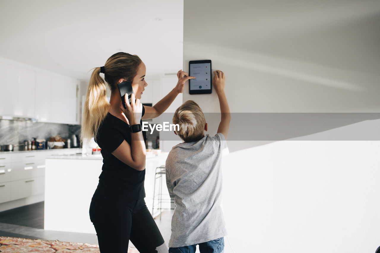 Teenage girl talking on phone using digital tablet by brother standing at home