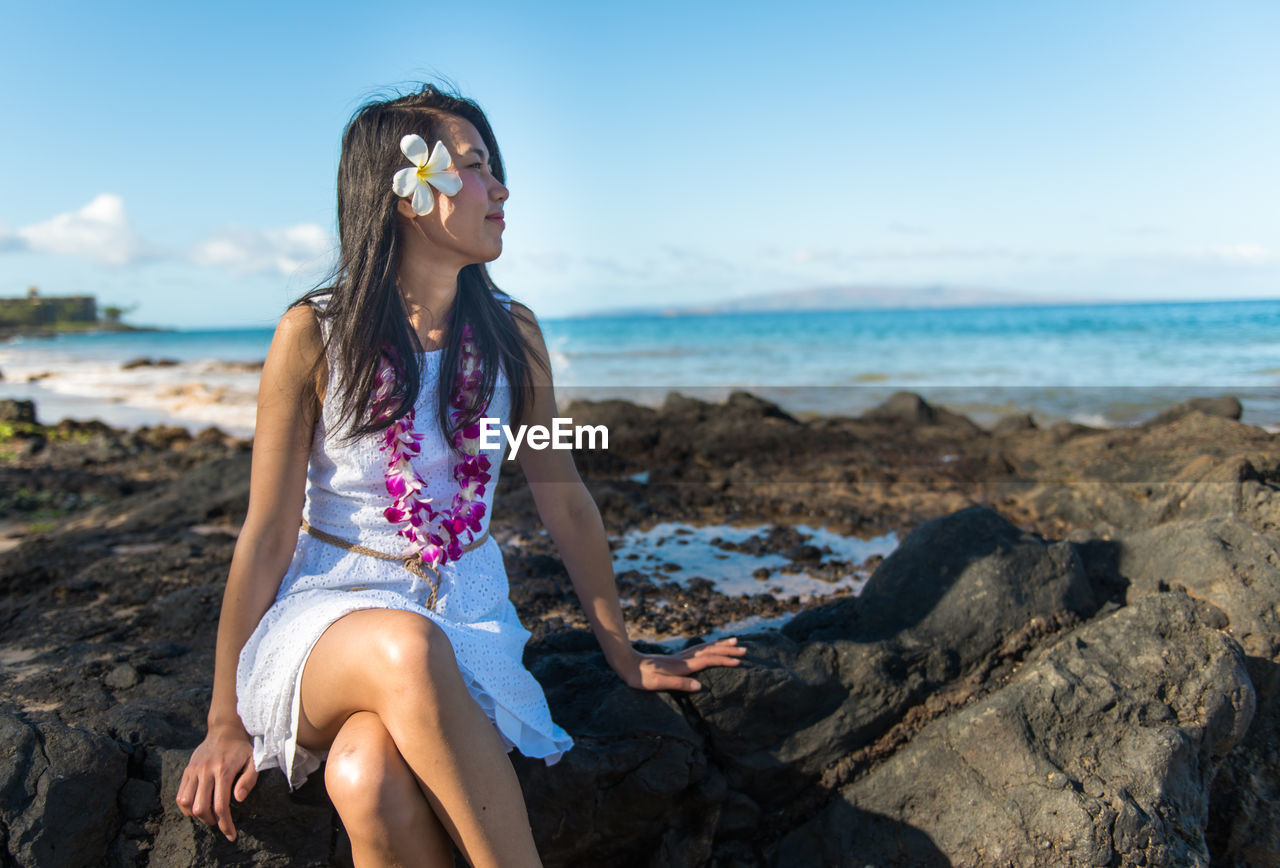 YOUNG WOMAN SITTING ON ROCK AT BEACH