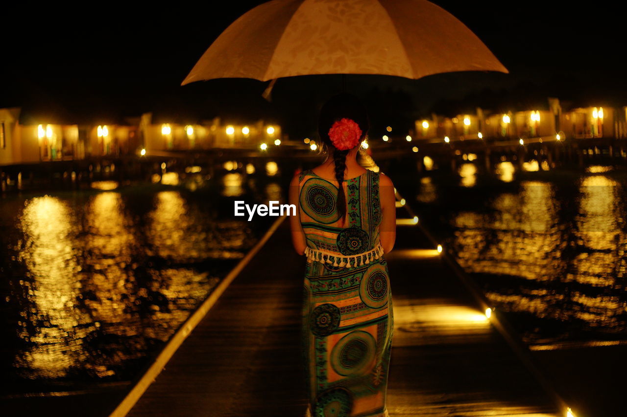 Rear view of woman with umbrella standing on pier over lake at night