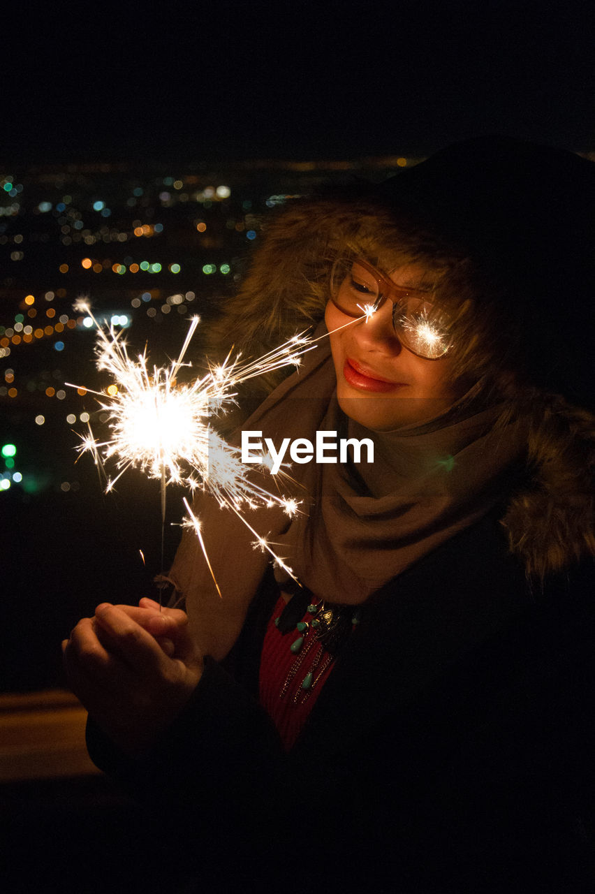 Smiling woman holding illuminated sparkler by window at night