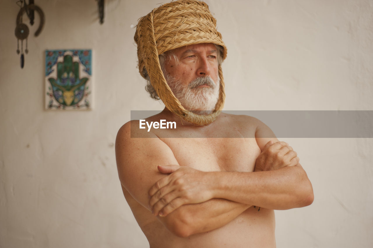 Shirtless man wearing basket while standing against wall