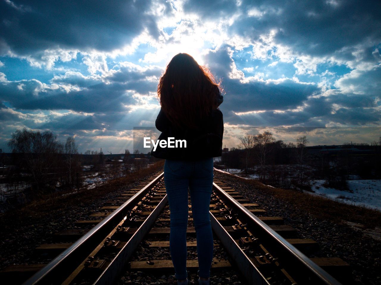 REAR VIEW OF WOMAN STANDING BY RAILROAD TRACKS AGAINST SKY