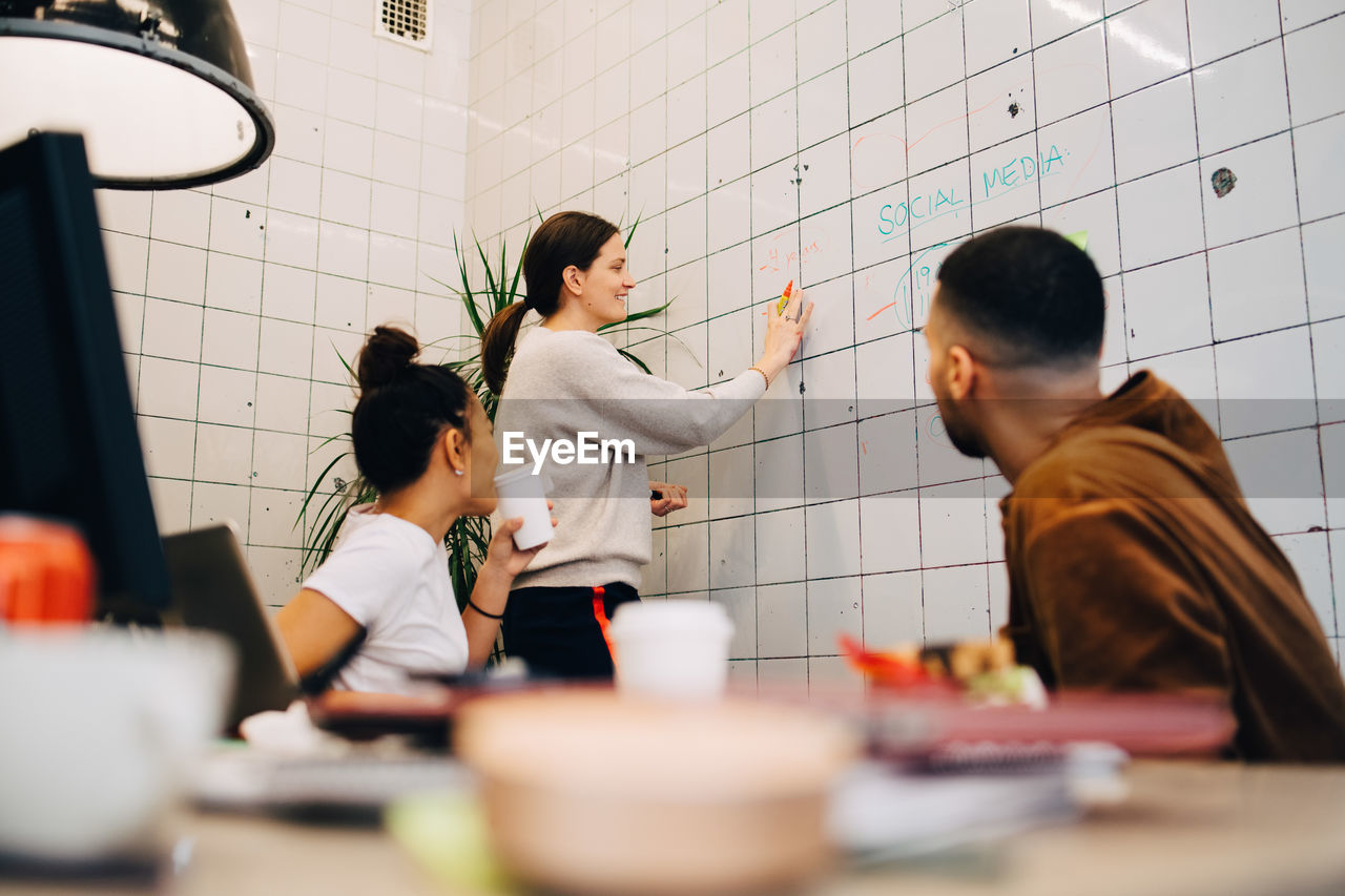 Smiling businesswoman discussing strategy on wall with colleagues at small creative office