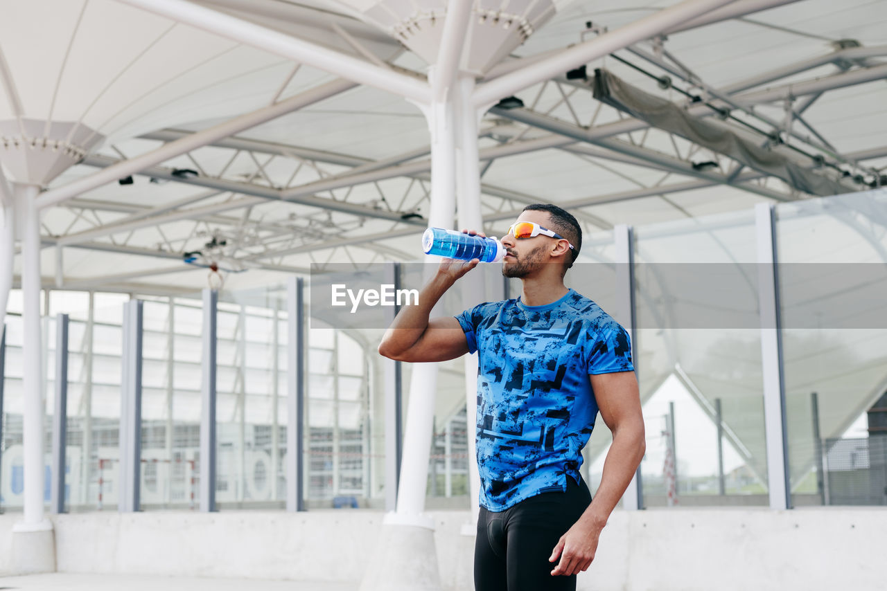 Athletic man with sports clothes and sunglasses standing and drinking water on the running track