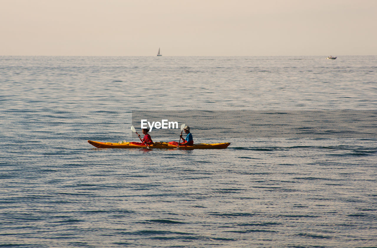 water, nautical vessel, transportation, sea, sky, boat, nature, sea kayak, mode of transportation, men, vehicle, kayak, boating, two people, horizon over water, adult, adventure, leisure activity, horizon, travel, oar, lifestyles, outdoors, togetherness, sports, beauty in nature, motion, day, holiday, kayaking, vacation, trip, sunset, scenics - nature, paddle, clear sky, watercraft, recreation, sports equipment, copy space, tranquility, women, environment, on the move, occupation, orange color, sitting, rippled, waterfront, activity, cooperation, teamwork, outdoor pursuit, tranquil scene, life jacket