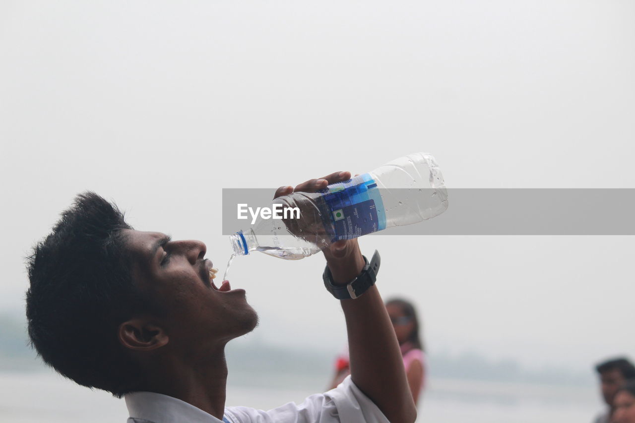 CLOSE-UP OF WOMAN HOLDING BOTTLE BY WATER AGAINST SKY