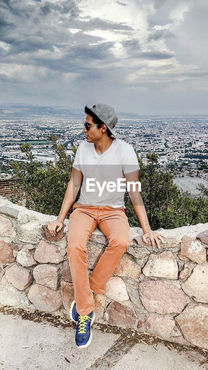 Man sitting on retaining wall against cityscape