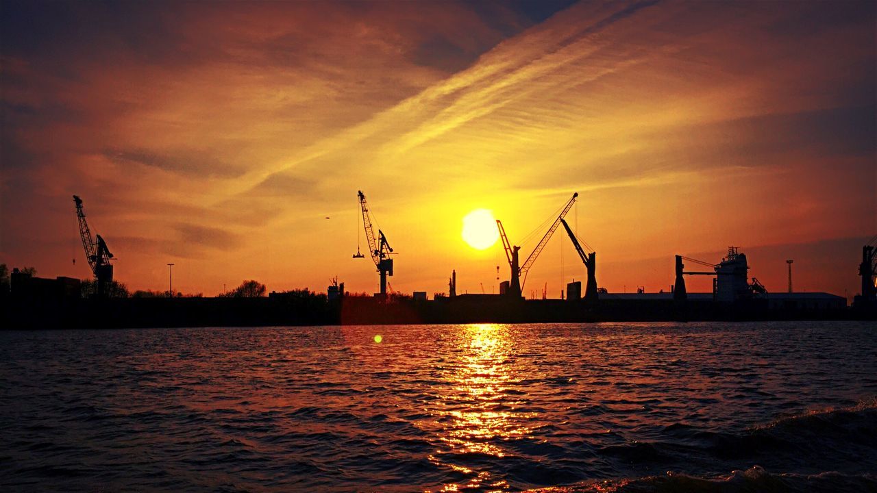 Silhouette cranes at harbor against orange cloudy sky
