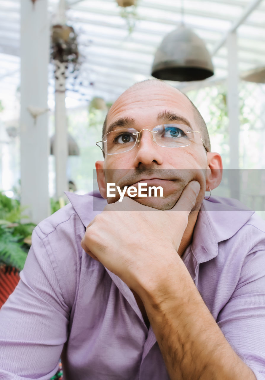 Attractive adult man with glasses holds his chin with the hand and looking up in a pensive mood.