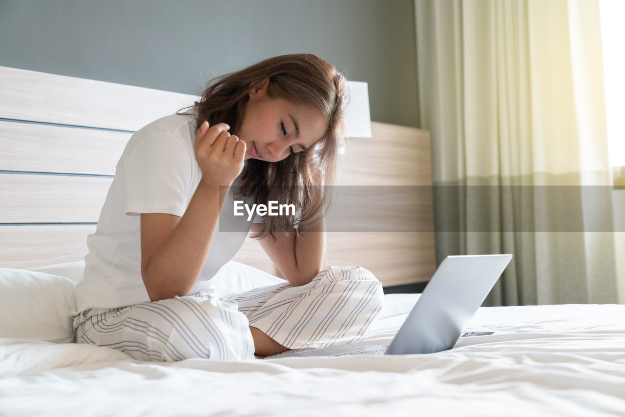 YOUNG WOMAN USING PHONE WHILE SITTING ON BED
