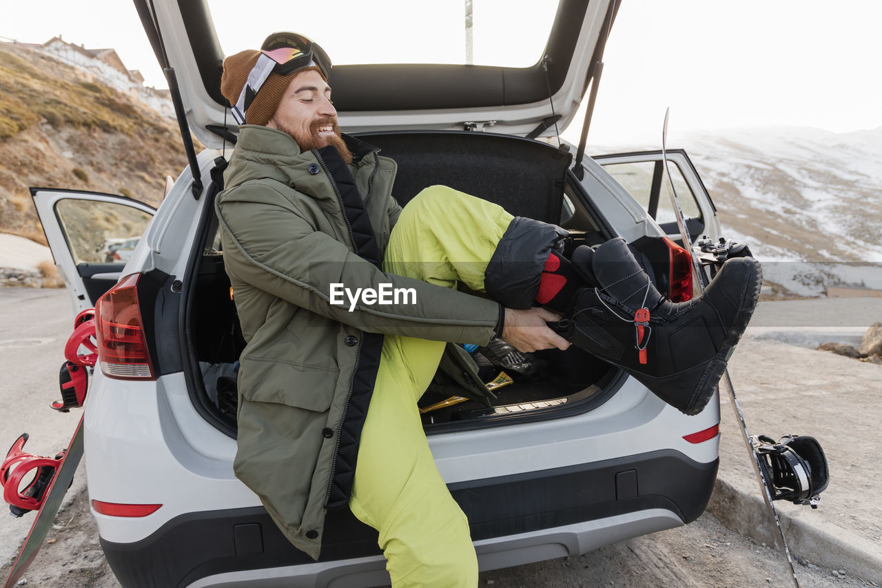 Smiling young man in car trunk putting on ski boots