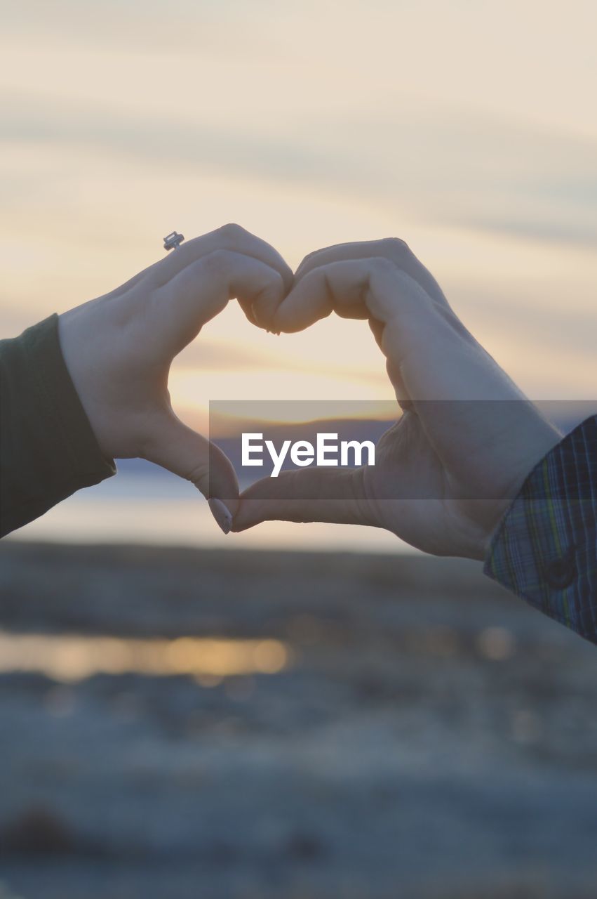 Cropped hands of woman making heart shape against sky during sunset