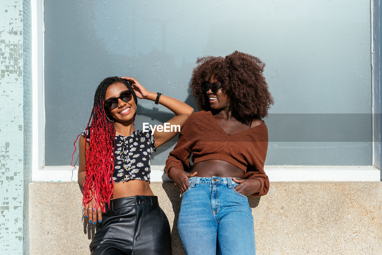 Black sisters in sunglasses standing on pavement and leaning on building wall while chilling in daylight