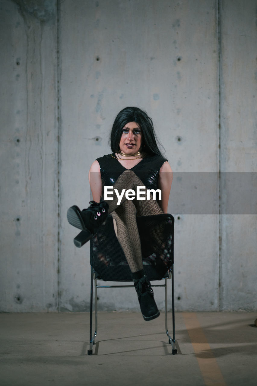 Full body of stylish drag queen wearing black garment and wig looking at camera while sitting on chair in studio