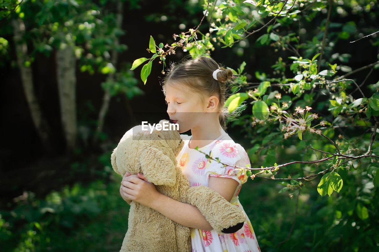 Cute european kid girl in dress with soft big toy dog in backyard, in park summer