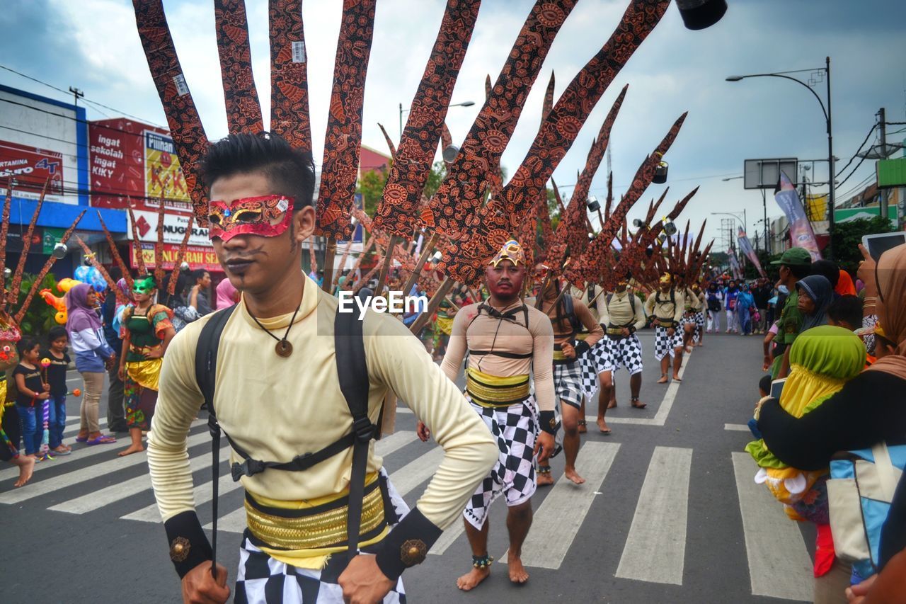 PEOPLE WALKING ON STREET AMIDST TREES IN CITY AGAINST SKY