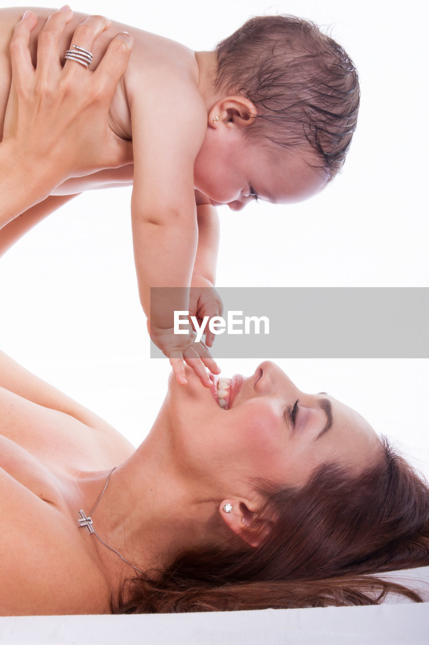 Happy mother playing with shirtless daughter while lifting her against white background