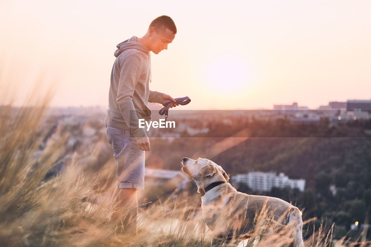 Side view of man with dog standing on field against sky during sunset
