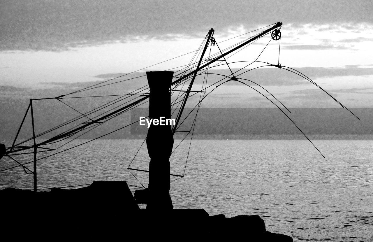 Silhouette fishing equipment on pole by sea against sky