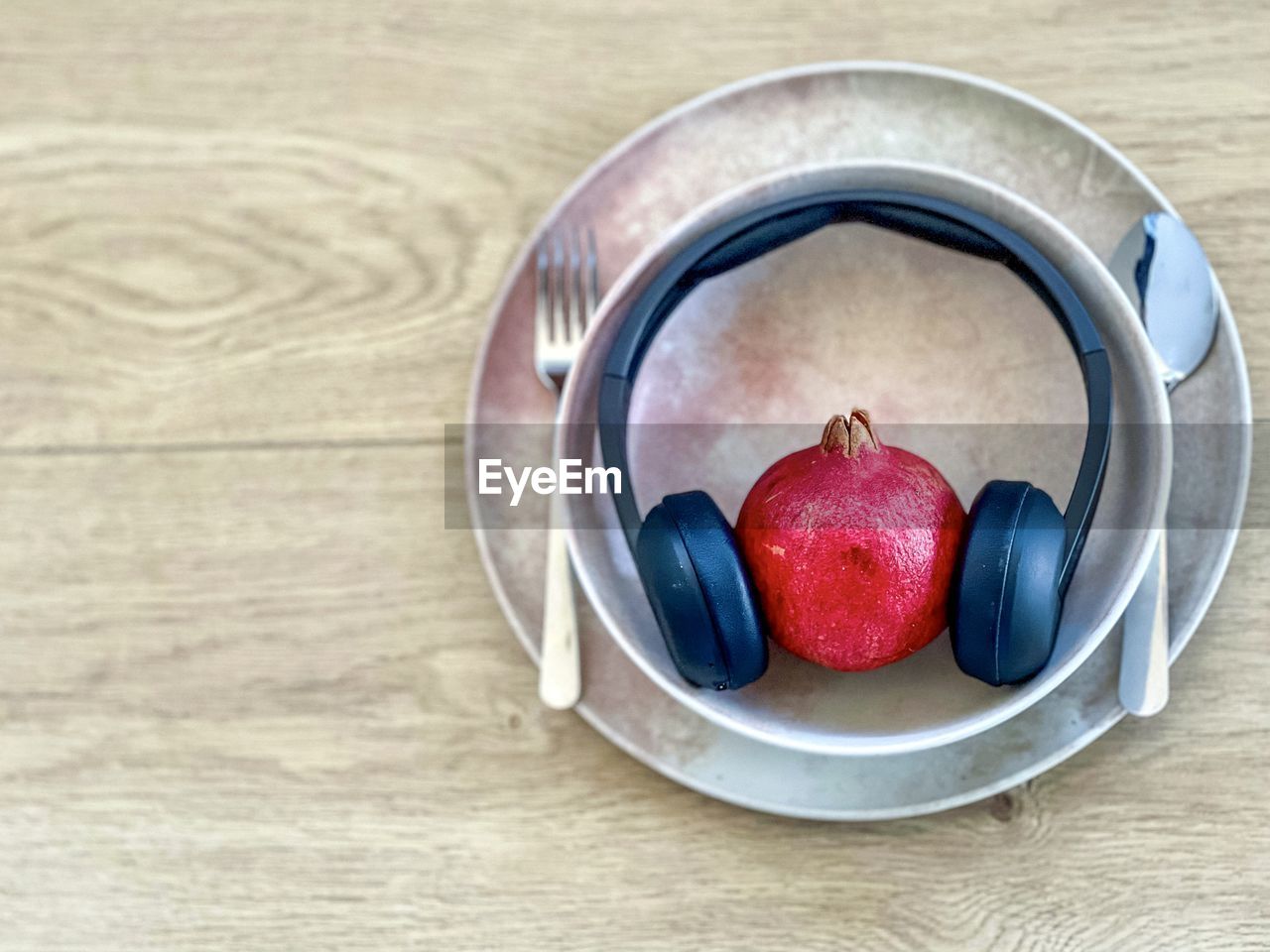 Conceptual shot of pomegranate and headphones in bowl on plate on table with cutlery.