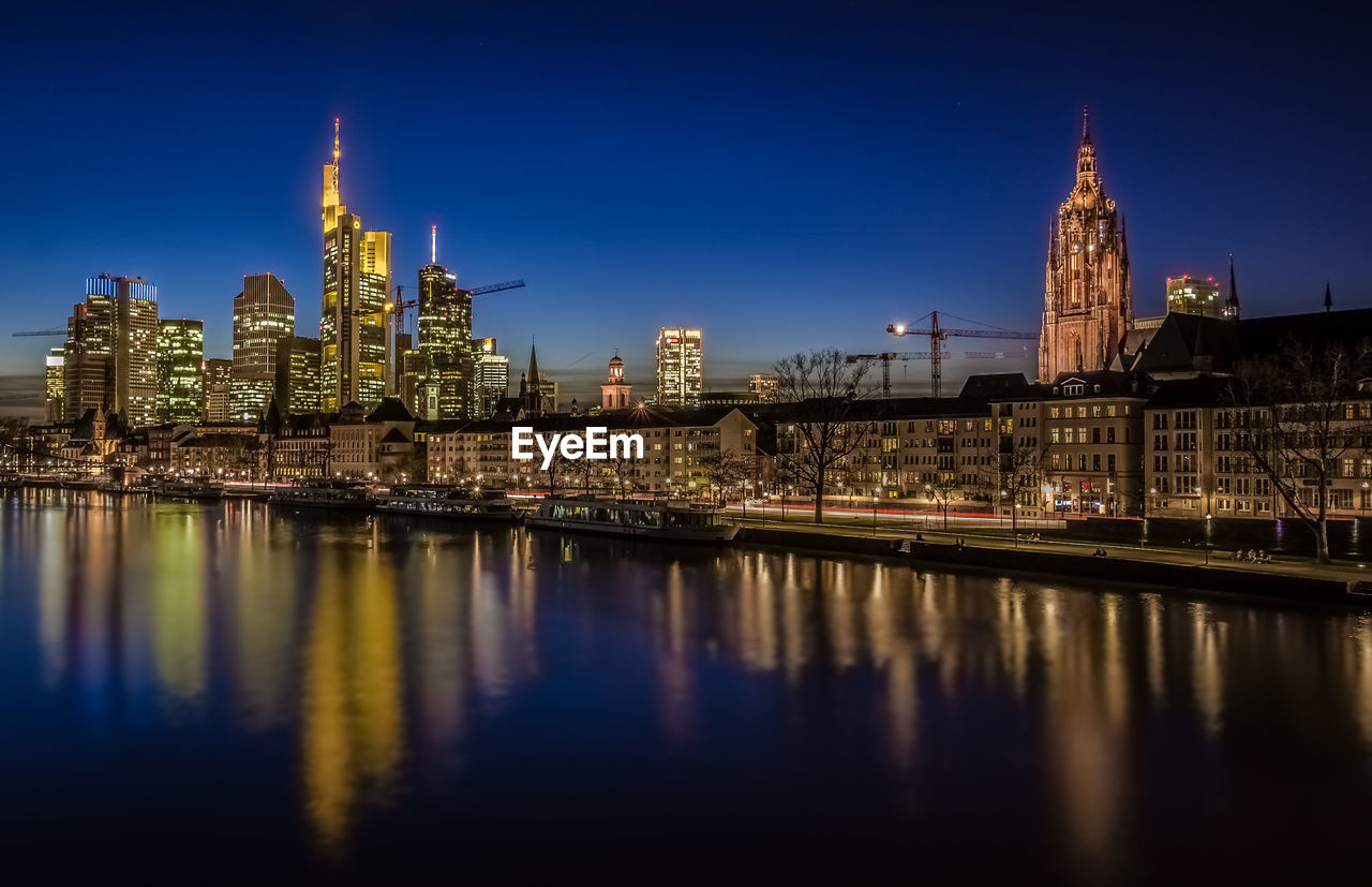 River by illuminated modern city against sky at dusk