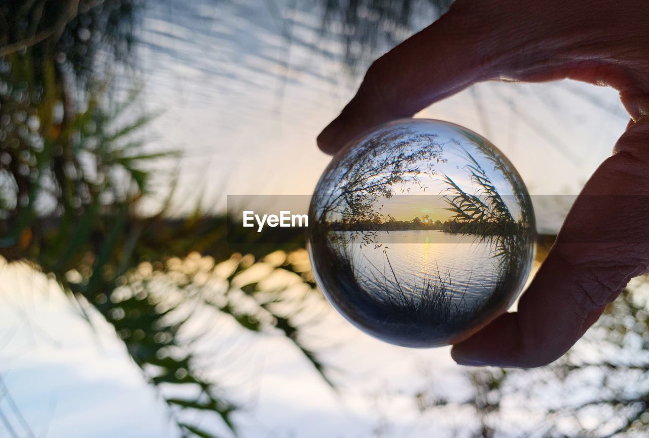 Cropped hand holding crystal ball with reflection on lake during sunset