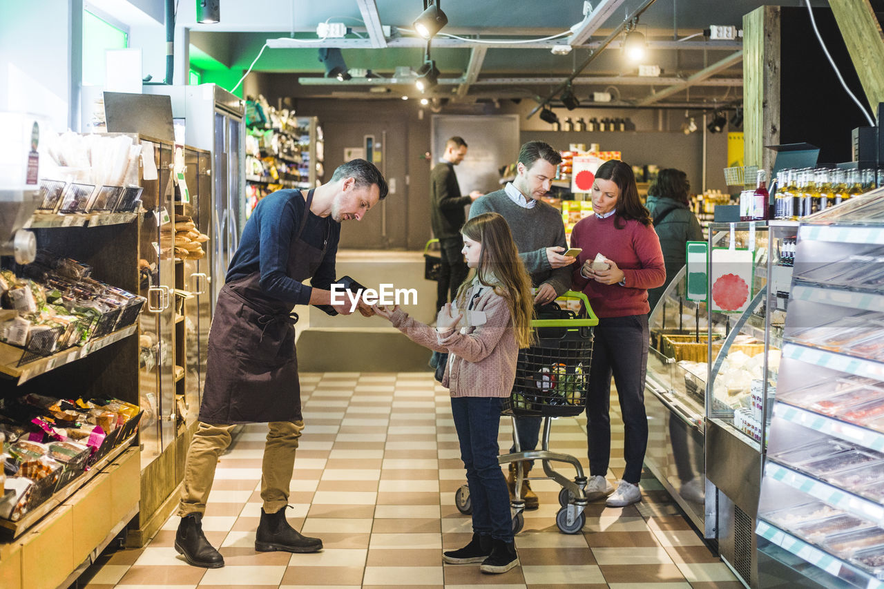 Store owner helping girl while couple discussing over mobile phone in supermarket