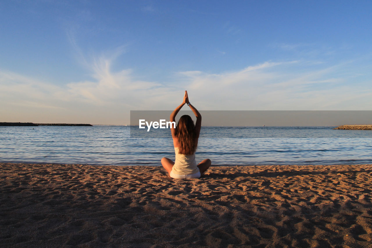 Rear view of woman practicing yoga on beach against sky at morning