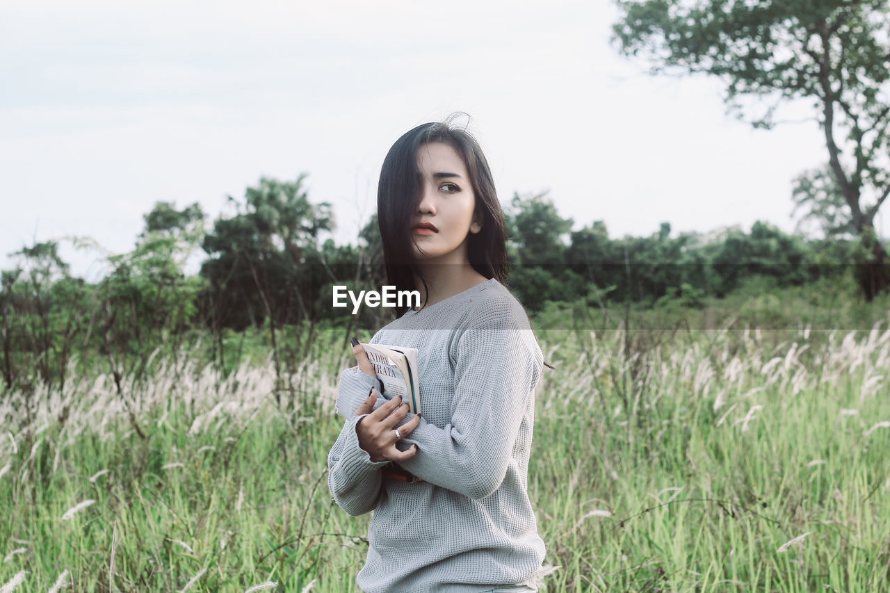 Thoughtful young woman holding book while standing on grassy land