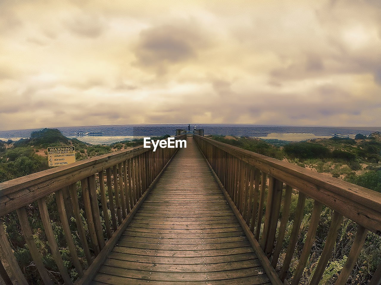 Wooden boardwalk leading towards sea against cloudy sky