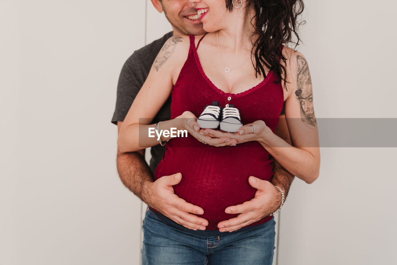 Midsection of pregnant couple holding shoes while standing against wall