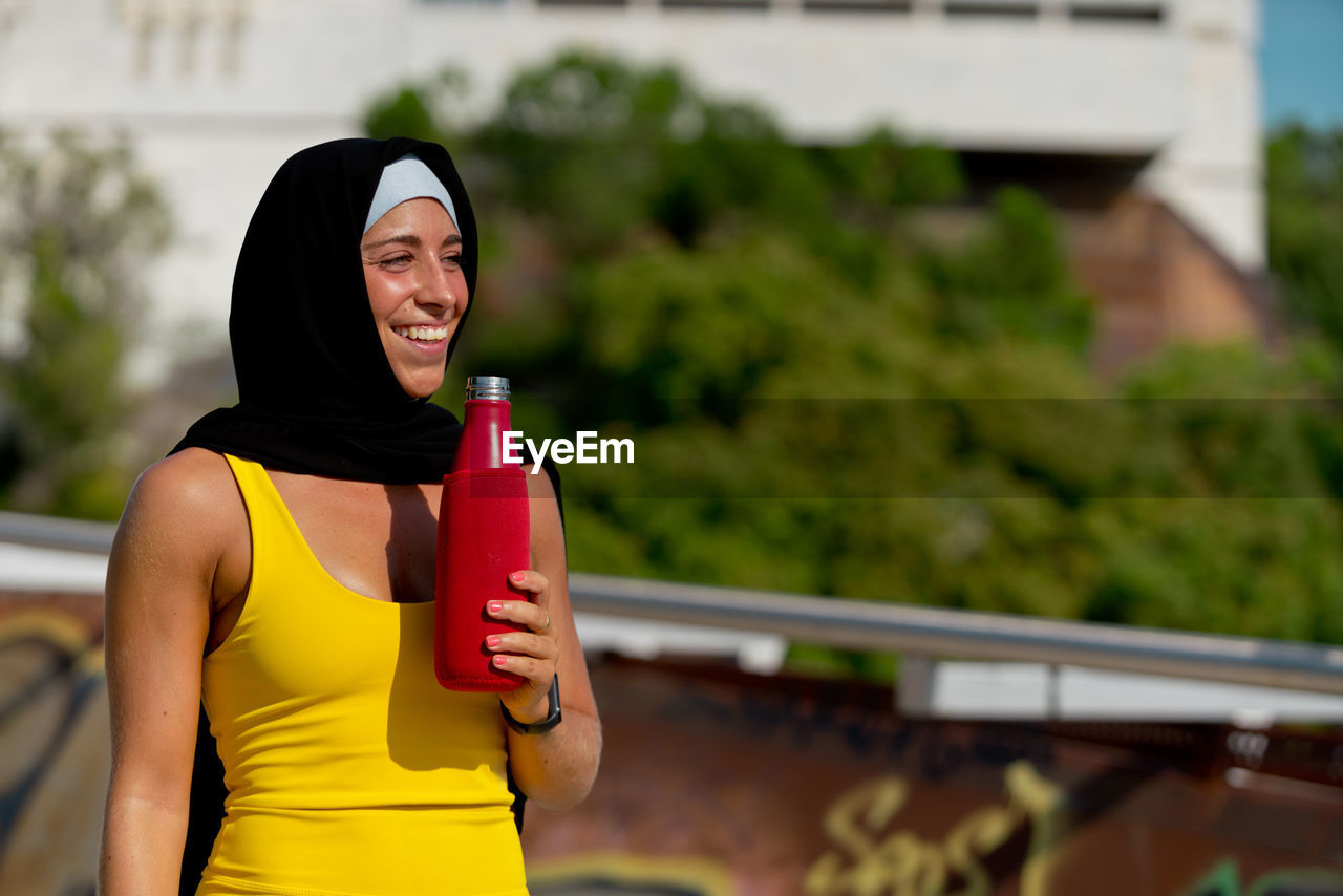 Smiling woman in hijab drinking water outdoors