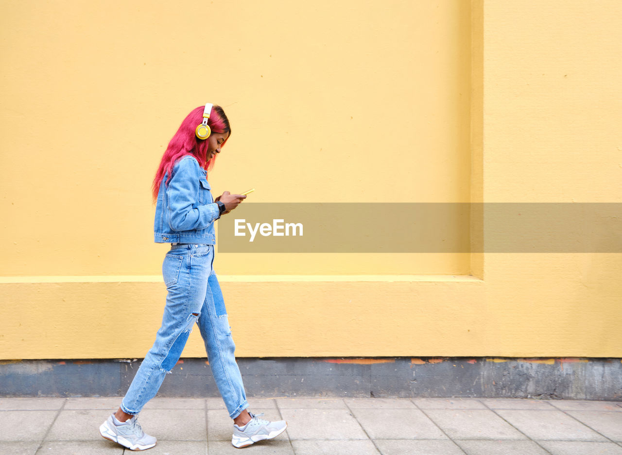 Happy young african american female hipster with pink dyed hair dressed in trendy denim outfit enjoying music through wireless headphones with smartphone while walking against yellow wall on street