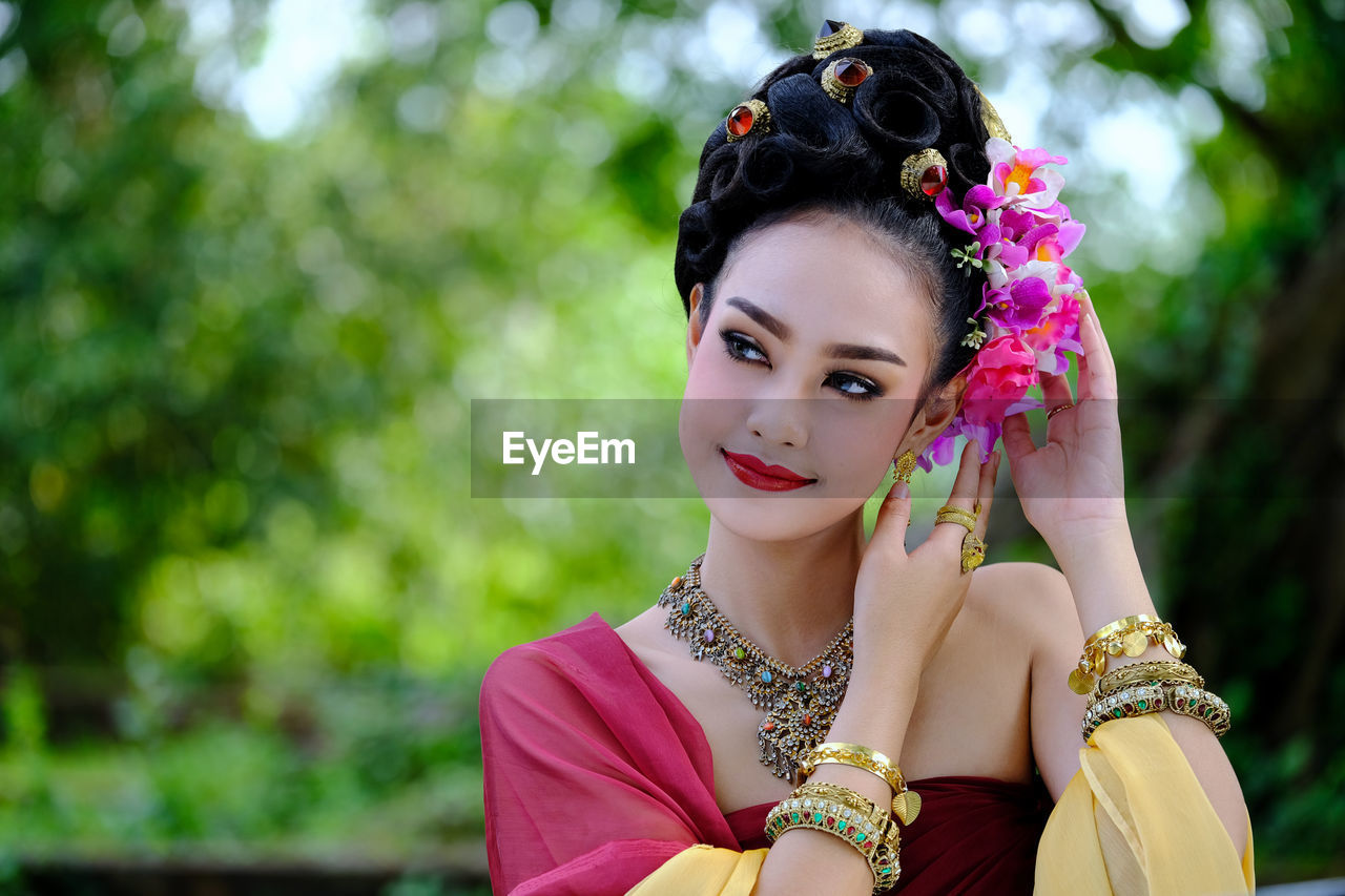 Close-up of thoughtful model in traditional clothing wearing flowers against trees