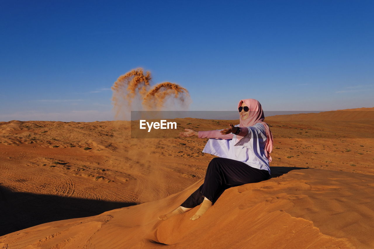 Full length of happy woman wearing hijab throwing sand on desert against sky