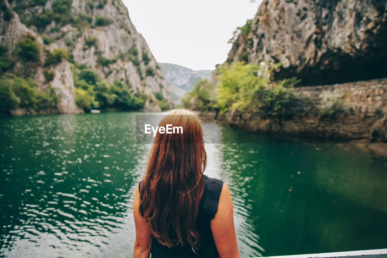 REAR VIEW OF WOMAN LOOKING AT LAKE BY MOUNTAIN