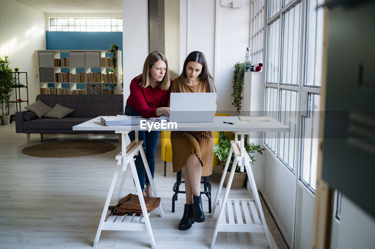 Businesswoman leaning by colleague using laptop at desk in office