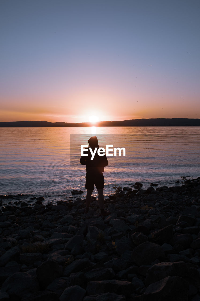 Man standing on rock at lakeshore against sky during sunset