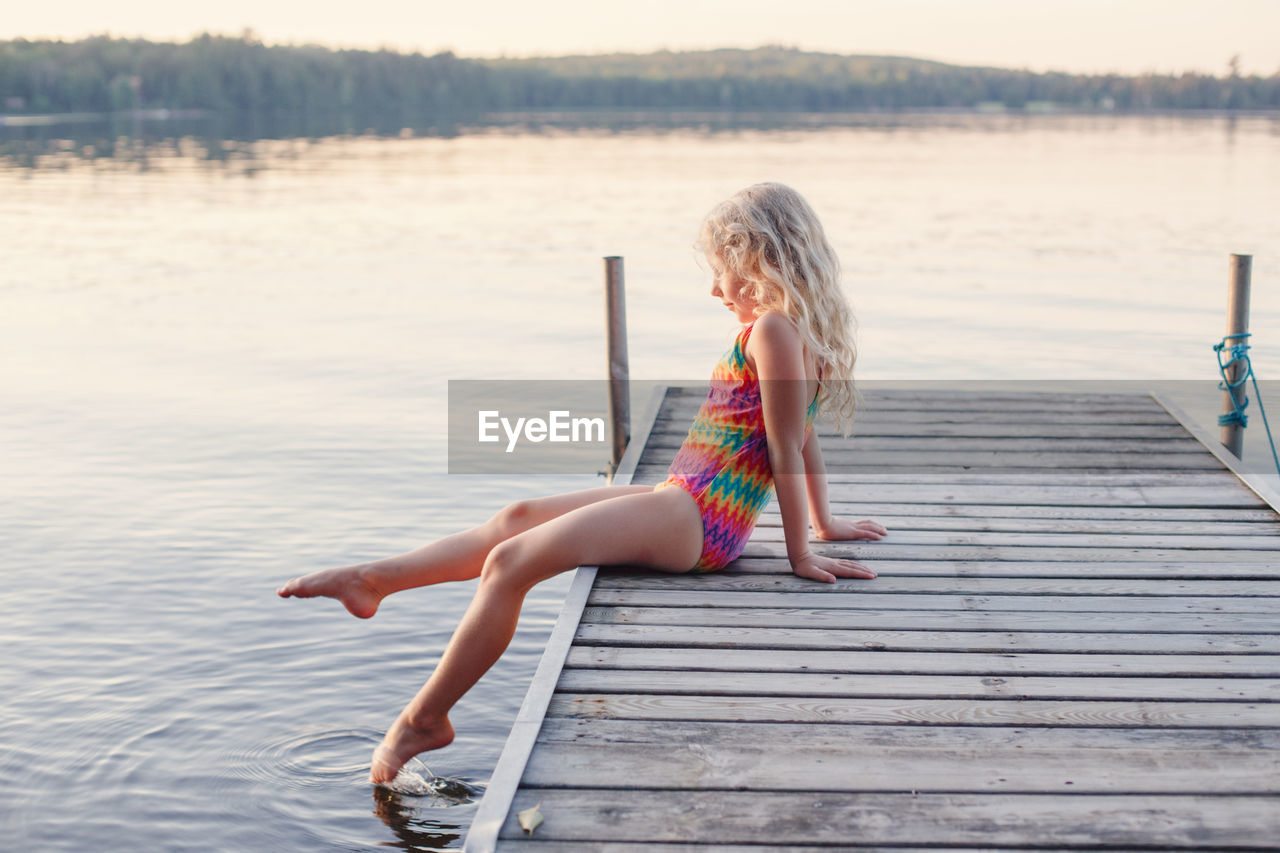 Caucasian blonde girl child sitting on wooden dock pier by lake. pensive kid in swimsuit splashing 