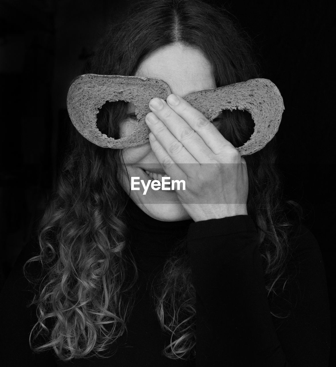 Portrait of woman covering eyes with bread slices against black background