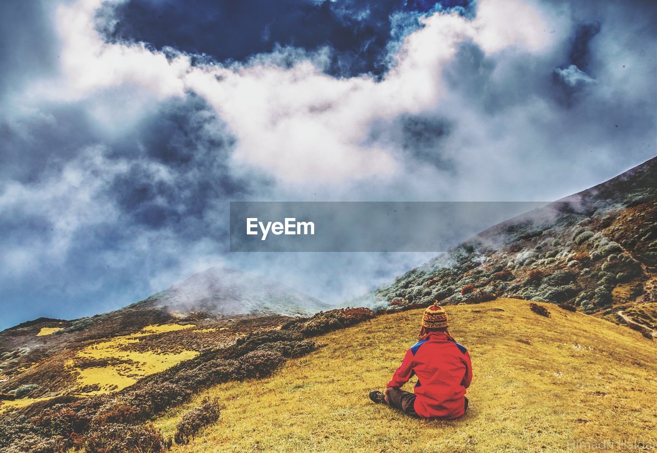 Rear view of man sitting on mountain against cloudy sky