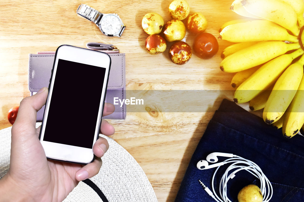 Cropped hand holding smart phone by fruits and personal accessories on table