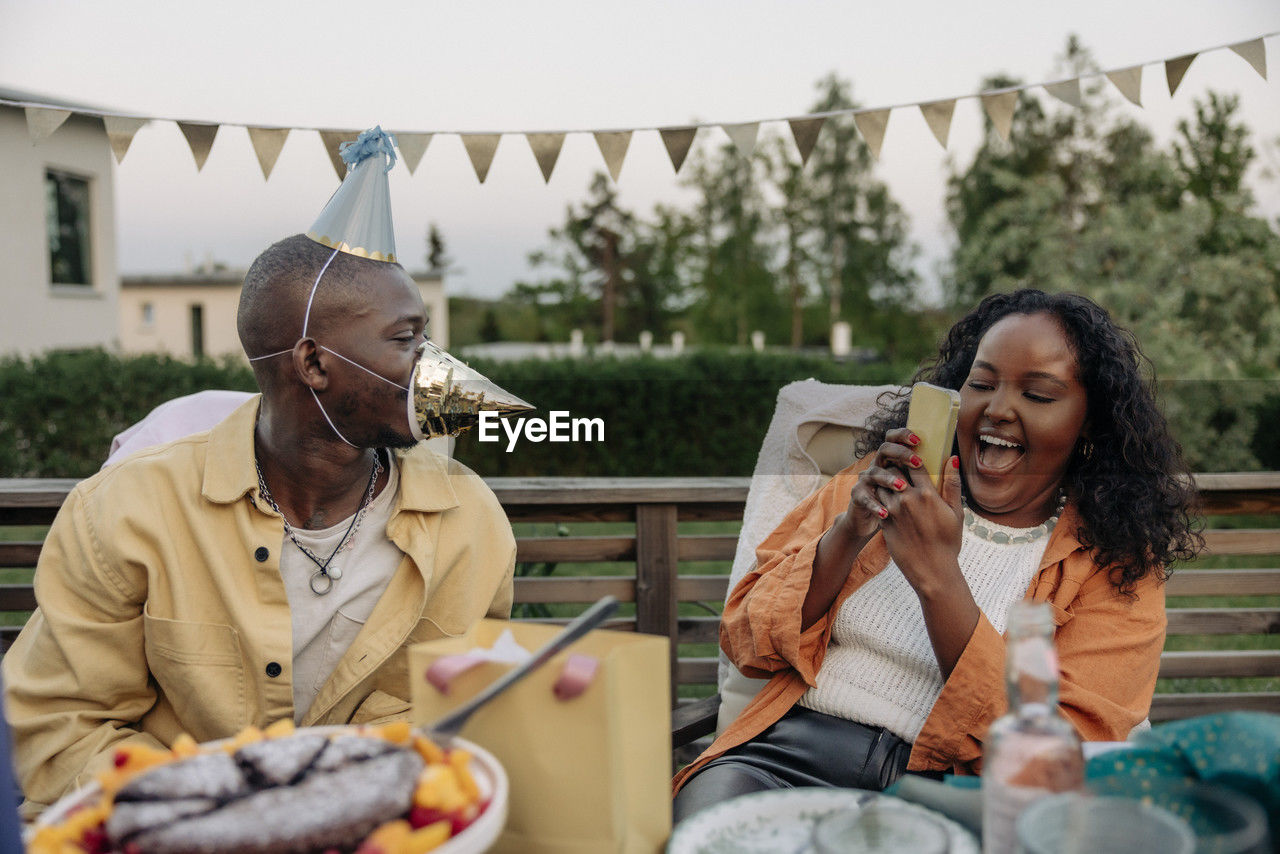 Happy woman photographing male friend wearing party hats while sitting in back yard