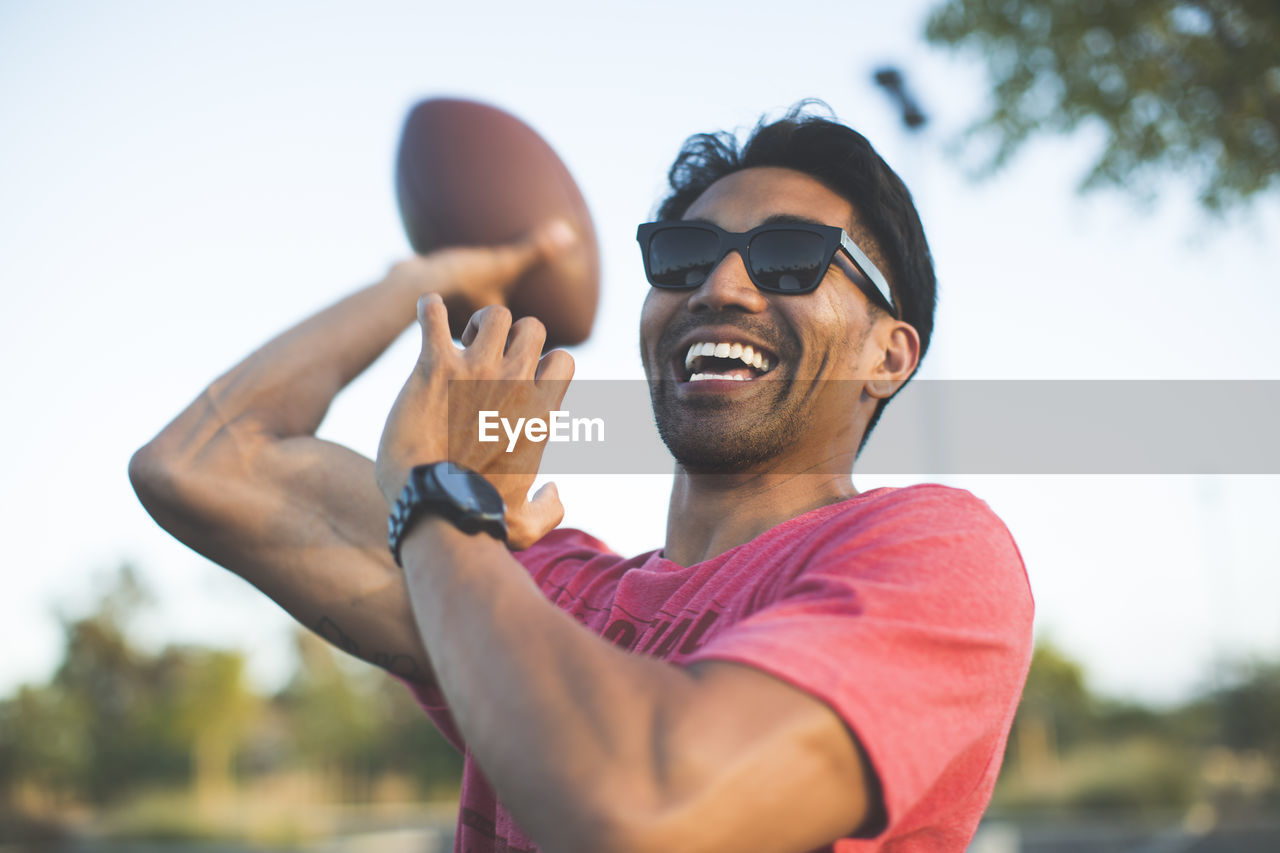 Cheerful man in sunglasses playing with american football against sky