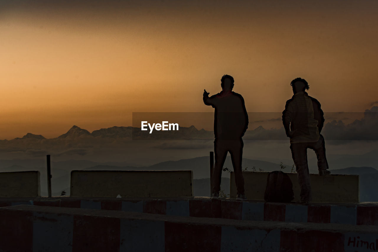 REAR VIEW OF MEN STANDING ON MOUNTAIN AGAINST SKY DURING SUNSET