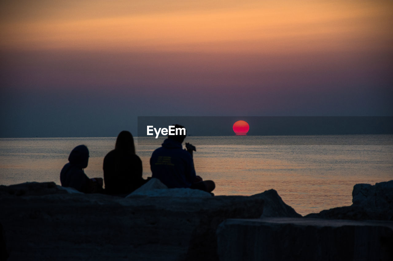 PEOPLE SITTING ON SEA SHORE AGAINST SUNSET SKY