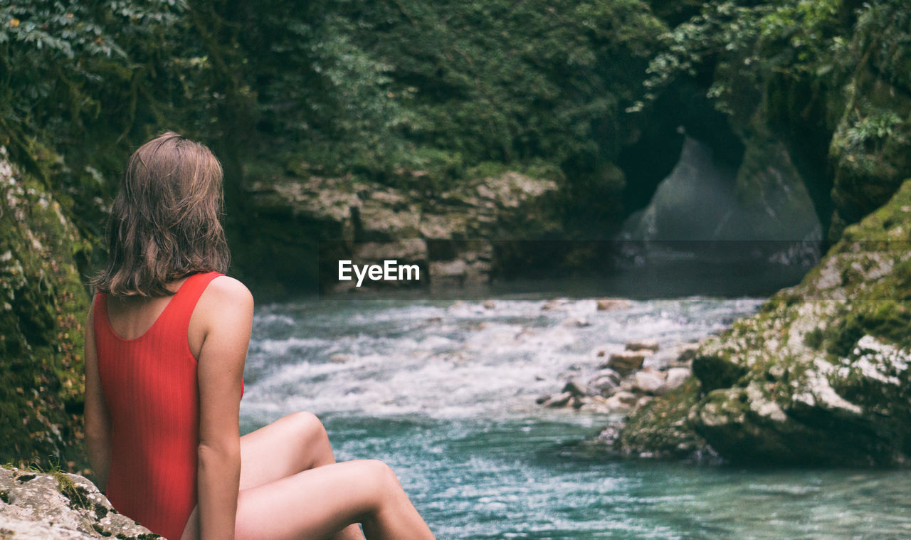 Rear view of woman sitting by river on rock at forest